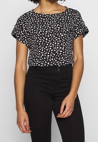 Czarna bluzka z krótkim rękawem w białe rozproszone kropki, okrągły dekolt i delikatne marszczenie. W połączeniu z dopasowanymi czarnymi spodniami.