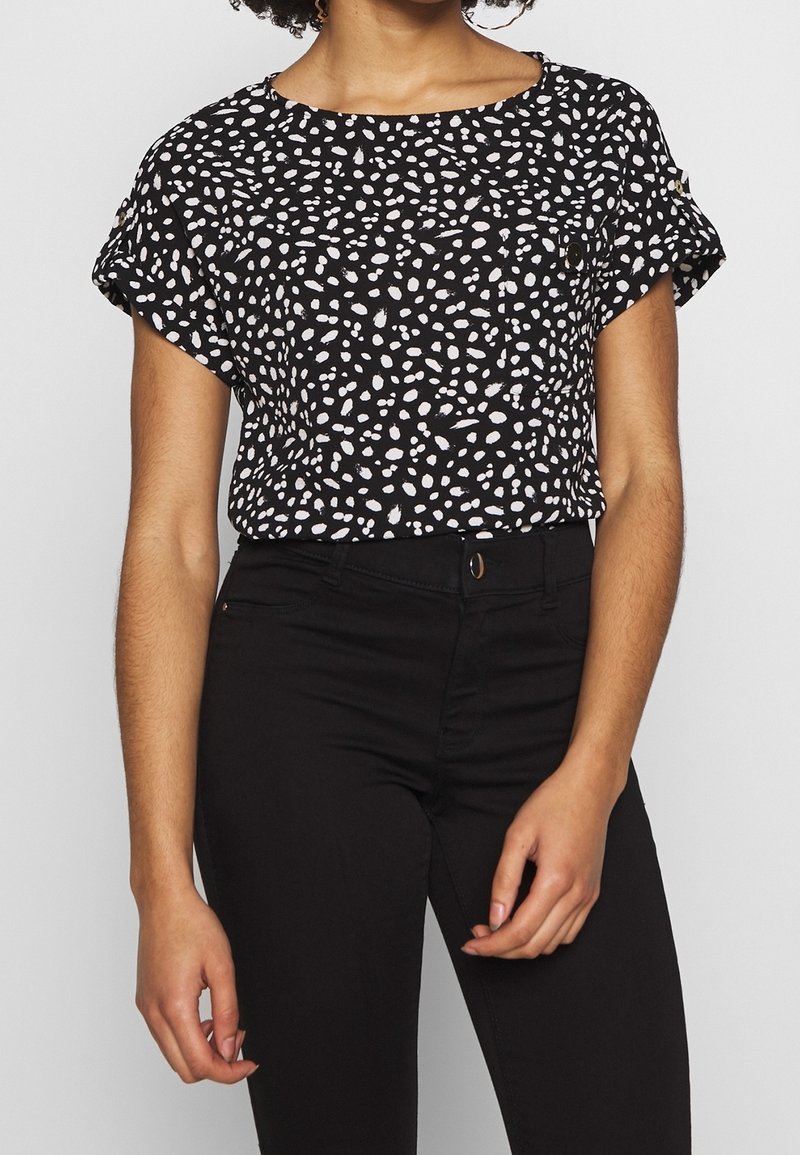 Czarna bluzka z krótkim rękawem w białe rozproszone kropki, okrągły dekolt i delikatne marszczenie. W połączeniu z dopasowanymi czarnymi spodniami.