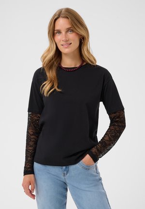 Femme aux longs cheveux blonds porte une chemise noire à manches longues en dentelle, un jean bleu et un collier de perles rouge foncé, souriant légèrement.