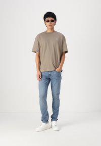 Svetlohnedé oversized tričko, modré džínsy a biele tenisky. Doplnky zahŕňajú štíhle čierne slnečné okuliare. Jednoduchý dizajn, voľný strih.