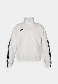 Giacca bianca con zip, caratterizzata da una texture a motivo, con accenti neri a tre strisce sulle maniche e un logo ricamato sul petto.