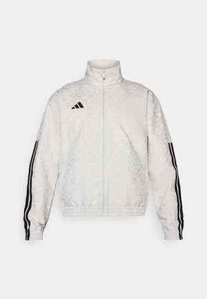 Hvit zip-up jakke med mønstret tekstur, med svarte tre-stripes aksenter på ermene og et brodert logo på brystet.