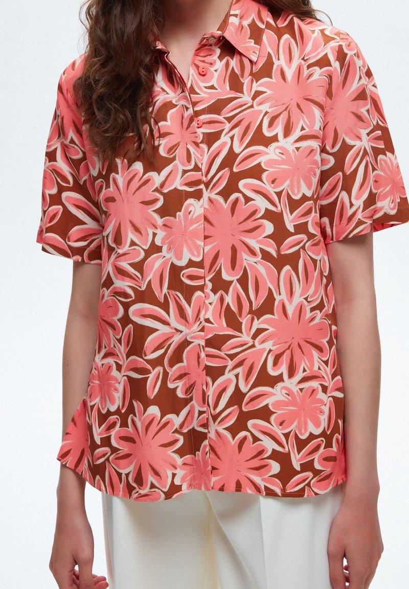 Femme portant une chemise à manches courtes boutonnée avec de grands motifs floraux roses sur un fond marron et un pantalon blanc.