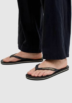 Feet wearing black Jack & Jones flip-flops with black pants against a plain light background.