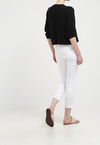 Mujer vestida con un suéter negro corto, pantalones blancos ajustados y sandalias planas beige, caminando de espaldas sobre un fondo blanco liso.