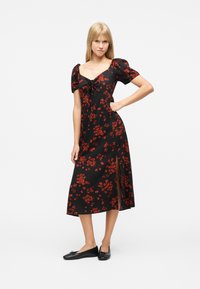 Femme portant une robe mi-longue noire à imprimé floral rouge, manches bouffantes, fente à l'avant, et des ballerines noires, debout devant un fond blanc.