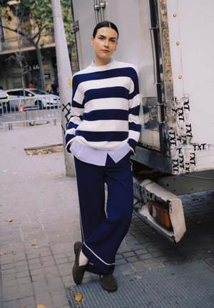 Ženska stoji na mestnem pločniku, oblečena v modro-belo črtasto pulover, modre široke hlače, z rokami v žepih, naslonjena na tovornjak.