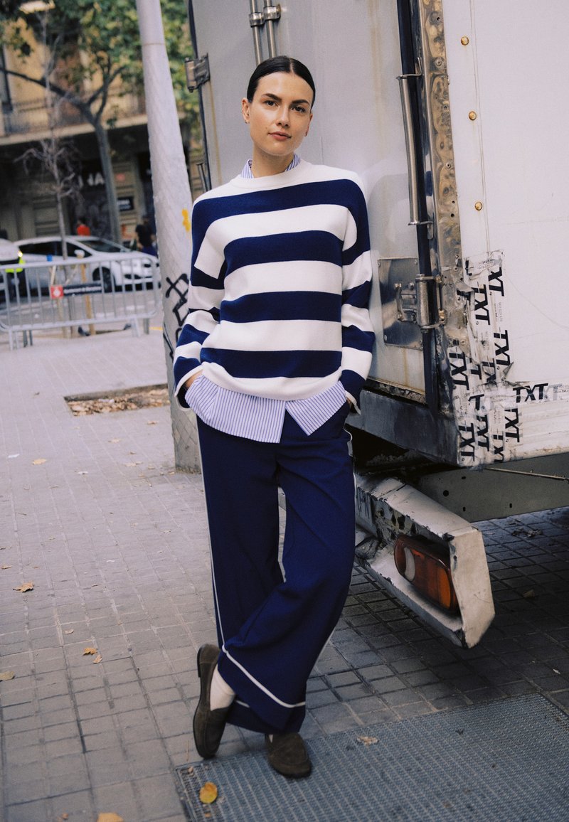 Mujer de pie en la acera de la ciudad, vistiendo un suéter a rayas navy y blanco, pantalones anchos navy, con las manos en los bolsillos, recostada contra un camión.