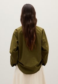 Persoon met lang, donker golvend haar, gekleed in een olijfgroene lange mouw shirt en een lichtbeige rok, staande tegen een effen lichte achtergrond.
