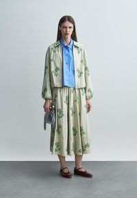 Femeie îmbrăcată cu o jachetă crem și fustă cu imprimeu floral verde, cămașă albastră cu nasturi, pantofi Mary Jane burgundy, ținând o geantă mică argintie.