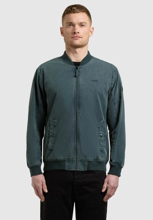 Muž v tmavozelenej bomber bunde na zips a čiernych nohaviciach stojaci pred jednoliatym svetlým pozadím.