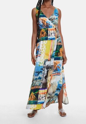 Femme portant une robe longue portefeuille sans manches avec des imprimés patchwork colorés et une fente latérale, associée à des sandales blanches.