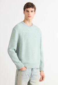 Világos menta zöld kötött pulóver, texturált mintával, kerek nyakkivágással, bordás mandzsettákkal és aljjal. Világoskék farmernadrággal viselve.