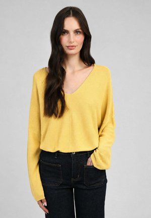 Femme aux longs cheveux foncés portant un pull jaune ample à col en V et un jean sombre taille haute, une main dans la poche, fond neutre.