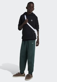 Svart hoodie med logotyp, gröna sweatpants, bruna sneakers och vit crossbody-väska. Tyget är mjukt; byxorna har en tapered passform.