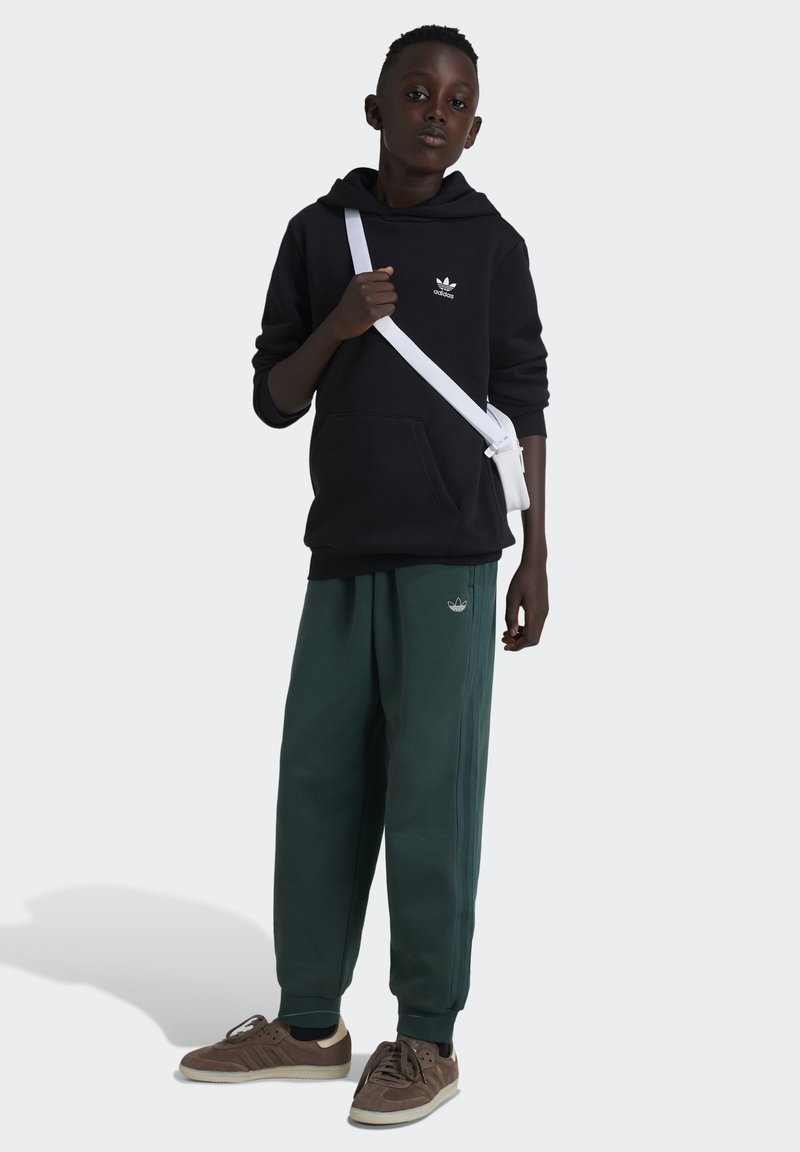 Svart hoodie med logotyp, gröna sweatpants, bruna sneakers och vit crossbody-väska. Tyget är mjukt; byxorna har en tapered passform.