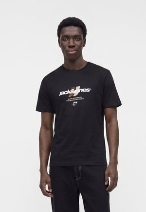 Ung mand står mod en ensfarvet lys baggrund, iført en sort "Jack & Jones" t-shirt og sorte jeans, og kigger mod kameraet.