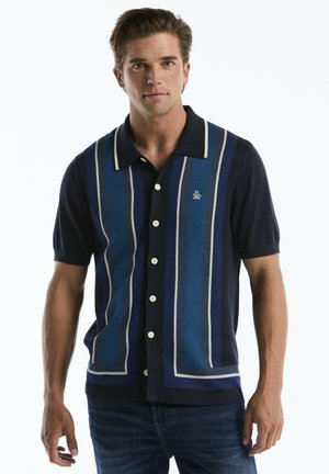 Mann trägt ein dunkelblaues kurzärmeliges Hemd mit blauen und weißen vertikalen Streifen und dunkle Jeans, steht vor einem weißen Hintergrund.