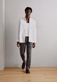 White striped button-up shirt over a white tank top, paired with gray trousers and black shoes, standing on a light wooden floor.