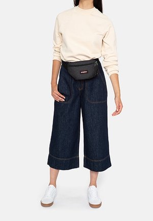 Felpa beige, culotte in denim scuro con gambe ampie e orli risvoltati, abbinati a sneaker bianche e una marsupio nero.