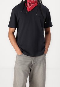 Camiseta de algodón azul marino de manga corta, corte relajado, pequeño logo en el pecho, combinada con pantalones de mezclilla gris claro. Un pañuelo añade un toque de rojo.