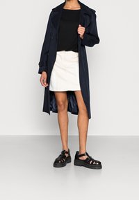Gabardina navy con diseño de doble botonadura, parte superior negra, falda crema y sandalias negras de plataforma. Tejido suave con un corte ajustado.