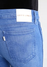 Siniset farkut, joissa on keskipäivän nousu ja takataskut, sekä valkoinen etiketti, jossa lukee "LEVI'S LINE 8" vyötäröllä. Kankaan tekstuuri on puuvillaa.