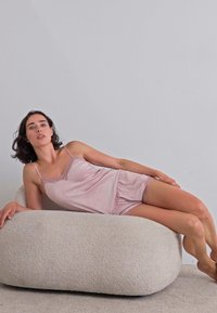 En ung kvinna i en rosa satinpyjamas ligger avslappnat på en mjuk, rund, beige ottoman mot en ljusgrå vägg.