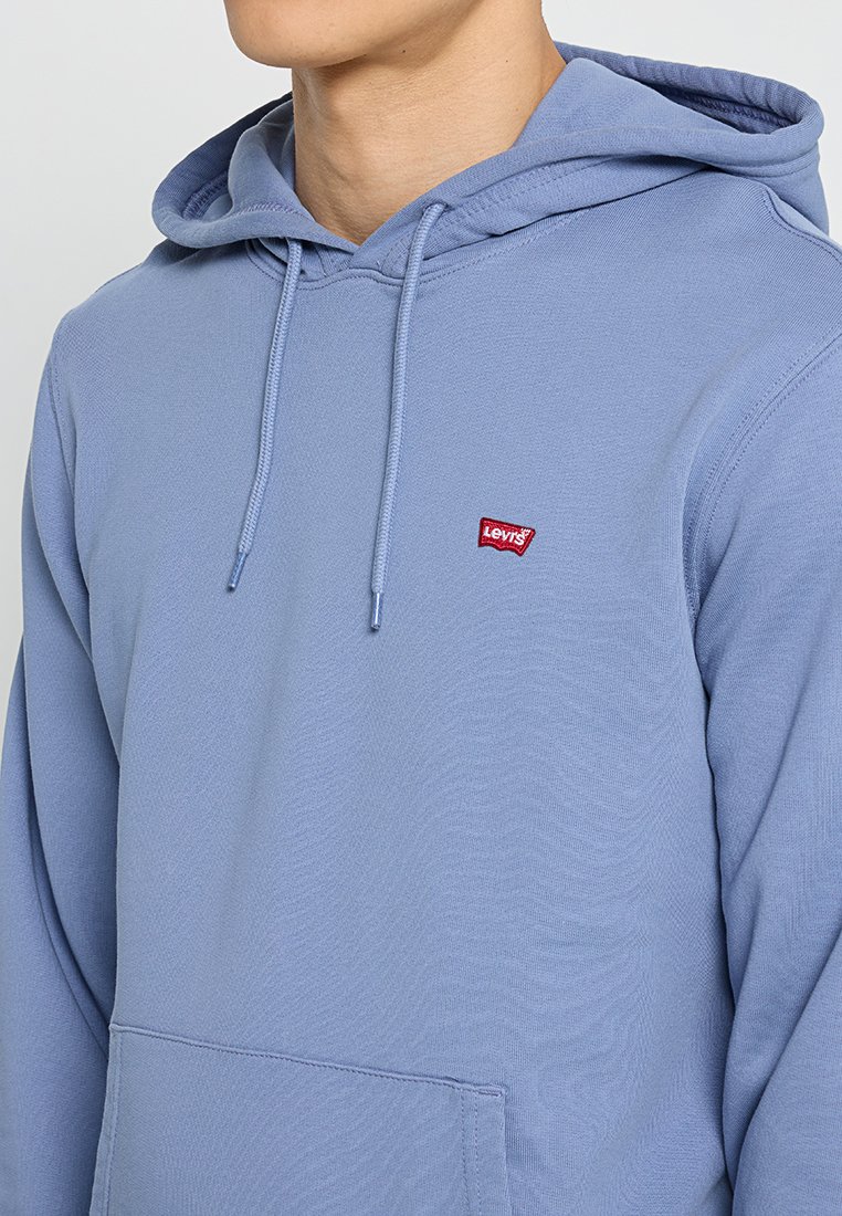 Hombre que lleva una sudadera ligera de Levi's azul claro con cordones visibles, mostrando parcialmente el torso y el cuello frente a un fondo liso.