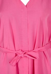 Różowa koszula z dekoltem w szpic, detalem w postaci wiązania w talii oraz marszczoną strukturą materiału. Materiał wydaje się lekki i gładki.