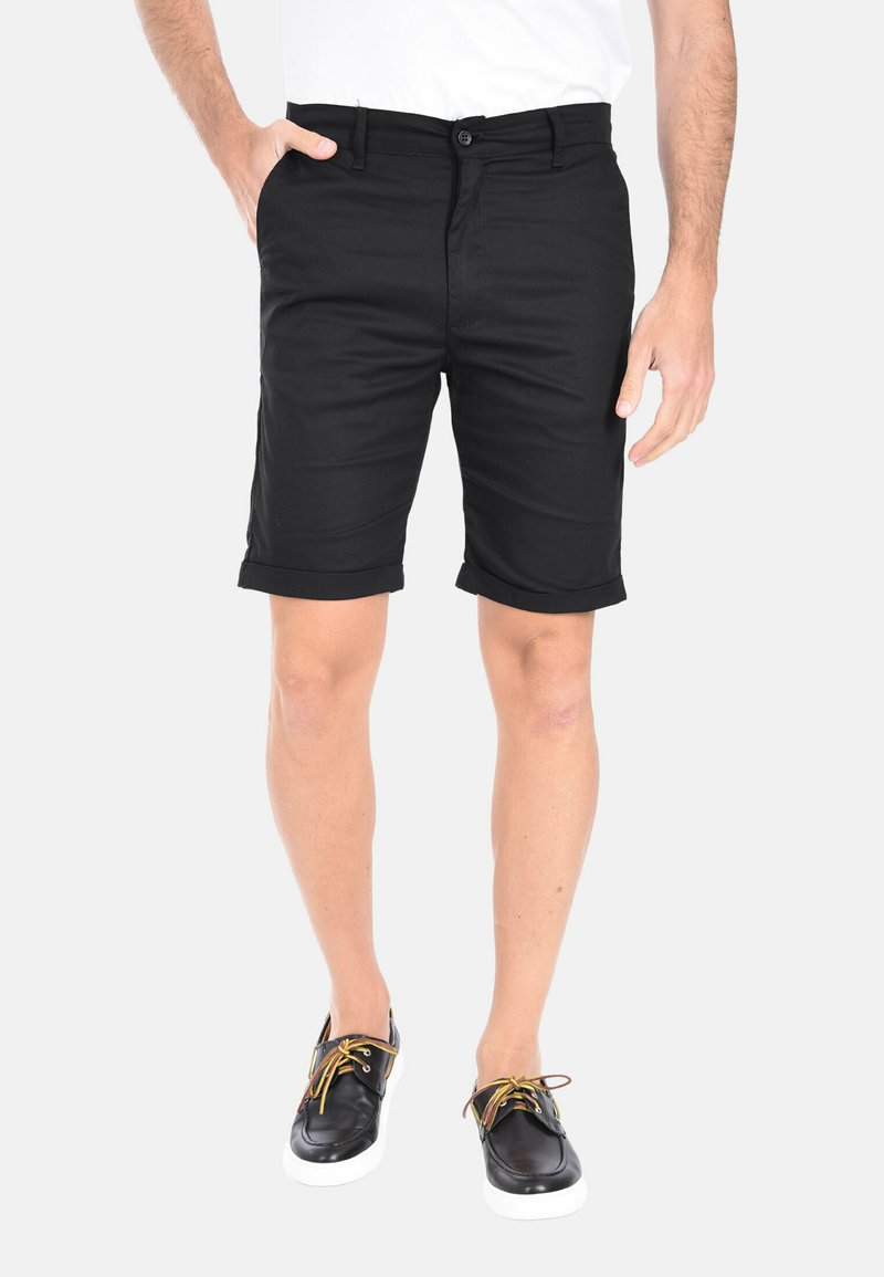 Pantaloni corti neri in cotone con una vestibilità sartoriale, dotati di chiusura con bottone, orli arrotolati e tasche laterali, abbinati a scarpe nere con lacci colorati.