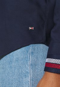 Marinblå kortärmad skjorta med en sidoflikslogotyp. Uppvikta ärmar med röda, vita och blåa randiga detaljer. Matchas med ljusblå jeans.