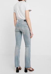 Mujer vestida con una camiseta ajustada de color gris claro, jeans rectos de mezclilla azul desvanecido y sandalias de tacón negras, de espaldas a la cámara.