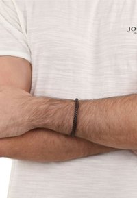 Braccialetto intrecciato in pelle marrone su un polso, indossato su una t-shirt chiara e texturizzata. I dettagli visibili includono il motivo dell'intreccio.