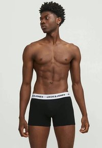 Μαύρα μπυφτερά σλιπ με λευκή επωνυμία στη μέση, προσφέροντας στενή εφαρμογή και μαλακή υφή. Το μοντέλο στέκεται με ίσια στάση, με τα χέρια χαλαρά.