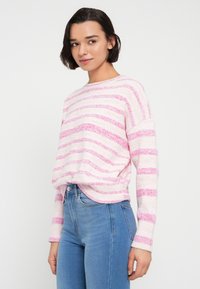 Ελαφρύ πλεκτό πουλόβερ με οριζόντιες ροζ και λευκές ρίγες, στρογγυλή λαιμόκοψη και μακριά μανίκια, συνδυασμένο με εφαρμοστά μπλε τζιν.
