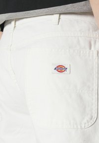 Hvide bomuldsbukser med en struktureret overflade, der har en baglomme og et fremtrædende logo patch i rød, gul og blå fra "Dickies".