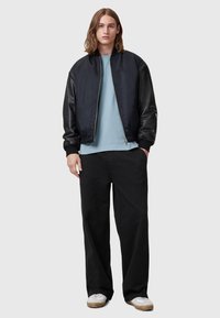 Giubbotto bomber nero con maniche in pelle, t-shirt azzurra chiara e pantaloni neri larghi. Abbinato a sneaker beige e una vestibilità rilassata.