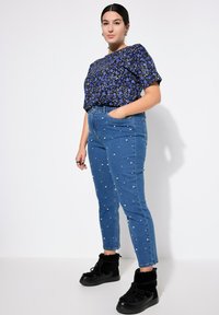 Top corto a maniche corte con stampa floreale blu abbinato a jeans in denim blu con decorazioni in perle bianche; modella che indossa stivaletti neri alla caviglia.