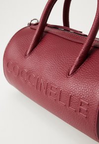 Burgundy leather handbag with textured finish, featuring two handles and embossed brand name "Coccinelle" on the side. Zip closure at top.