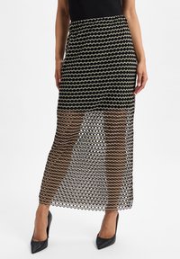 Maxirock aus schwarzem Mesh-Stoff mit wellenförmigen, ovalen Mustern; transparenter Mittelteil; elastischer Bund; kombiniert mit schwarzen High Heels.