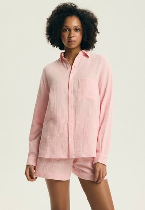 Kobieta o kręconych włosach, ubrana w jasnoróżową, teksturowaną koszulę na guziki z długim rękawem i pasujące do niej krótkie spodenki, stojąca na jednolitym tle.