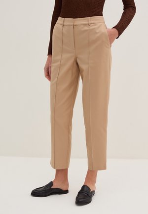 Beżowe, dopasowane spodnie do kostek noszone z brązowym, prążkowanym swetrem i czarnymi wsuwanymi loafersami, z jedną ręką w kieszeni.