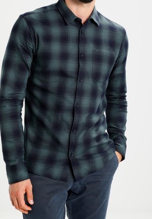 Man draagt een donkergroen en zwart geruit overhemd met knopen en lange mouwen, en donkerblauwe broek, handen in de zakken.