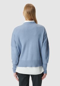 Pull en tricot bleu clair à coupe ample, avec une texture côtelée aux poignets et à l'ourlet, porté par-dessus une chemise blanche à col.