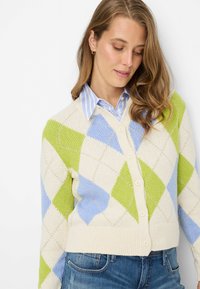 Cremefarbener Cardigan mit grün-blauem Argyle-Muster, Strickstruktur und Knöpfen vorne. Wird über einem blau gestreiften Hemd mit Kragen getragen.