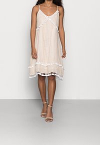 Nainen, joka on pukeutunut vaaleabeigeeseen, kuvioituun hihattomaan mekkoon, jossa on valkoista pom-pom-yskiä, ja vaaleabeigeisiin nilkkaketjullisiin korkokenkiin, seisoo yksinkertaista taustaa vasten.