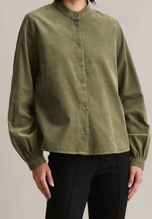 Kvinde iført en olivengrøn langærmet bluse med knapper og rynkede manchetter samt sorte, tætsiddende bukser, stående foran en neutral baggrund.