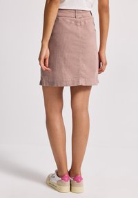 Kvinna som bär en dammig rosa kordskjorta och vita sneakers med rosa händelser, står mot en enkel vit bakgrund.