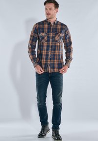 Karierte Flanellhemd in Blau- und Orangetönen, lange Ärmel, Knopfleiste, zwei Brusttaschen, kombiniert mit dunklen Jeans und schwarzen Stiefeln.